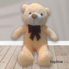 Large cream teddy bear plush sitting on floor with brown bow (product image).