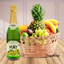 Fruit basket with pineapple, grapes, bananas and a bottle of sparkling drink labeled 'May'.