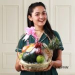Smiling woman holding a large fruit basket with pineapple, melon and assorted fresh fruit.