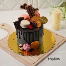 Chocolate drip cake topped with macarons, truffles and biscuit decorations on a gold board.